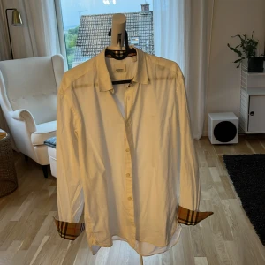Vit Burberry skjorta med rutiga detaljer - Säljer min burberry skjorta då jag inte använder den längre. Den är i size large och är i mycket bra skick bortsett från en liten fläck på ena axeln som inte märks om man inte letar efter den plus ett pyttelitet hål vid ärmen men den täcks över när du viker upp ärmen för att visa mönstret 