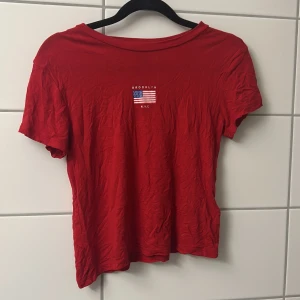 Röd t-shirt Brooklyn NYC - Röd t-shirt med kort ärm och tryck framtill med amerikansk flagga samt texten 'Brooklyn NYC'. Enkel och stilren modell med rund halsringning, perfekt för en avslappnad look.