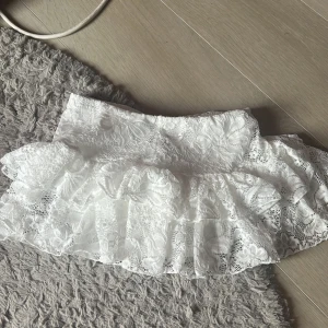 Vit spetskjol med volanger från SHEIN - Superfin vit kortkjol från SHEIN i spets med volanger. Kjolen har ett romantiskt uttryck med blommigt spetsmönster och flera lager volanger som ger extra fluff. Perfekt för dig som vill ha en söt och trendig look i sommar. Den har också inbyggda shorts som inte syns! 