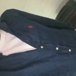Mörkblå skjorta från Polo Ralph Lauren - Snygg mörkblå skjorta från Polo Ralph Lauren med klassisk röd broderad logga på bröstet. Skjortan har långa ärmar, knappar framtill och är tillverkad i ett mjukt bomullstyg. Perfekt för dig som gillar stilrena och tidlösa plagg. Priset kan förhandlas 