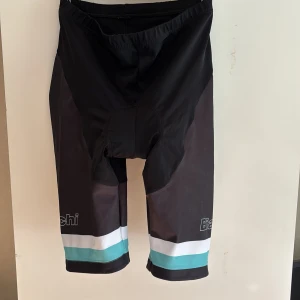 Bianchi cykelbyxor svart/turkos L - Snygga cykelbyxor från Bianchi i svart med vita och turkosa detaljer. Byxorna har vaddering för extra komfort och är tillverkade i en stretchig syntetblandning. Perfekta för dig som gillar att cykla och vill ha en sportig look.