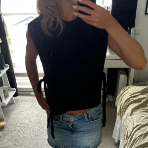 Svart väst med knyt i sidorna - Säljer en svart väst med ärmlös design och knytband i sidorna. Västen har en rak och lite boxig passform som ger en cool och avslappnad look. Perfekt att styla över en t-shirt eller skjorta för en trendig layering-effekt.
