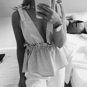 Randig blå och vit linnetopp med rosetter - Supersöt linnetopp med smala blå och vita ränder, djup v-ringning och knytrosetter på axlarna. Toppen har volangkant i midjan och är ärmlös, perfekt för varma dagar. Luftig och lätt i bomull, ger en fräsch och somrig vibe.