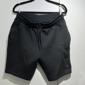 Svarta shorts från Gymshark - Svarta shorts från Gymshark med elastisk midja och snörning. Mjukt material och diskret logga på benet. Perfekta för gymmet eller chill dagar. Klassisk passform och stilren look.