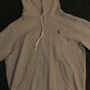 Grå hoodie från Polo Ralph Lauren - Snygg grå hoodie från Polo Ralph Lauren med klassisk broderad logga på bröstet. Tröjan har huva med snörning och en stor magficka. Perfekt för chill dagar och enkel att matcha med jeans eller joggers. Den kostar egentligen 2100 kr men mitt pris är 650 kr, vill bli av med den snabbt därför så lågt pris. Den är äkta och det finns även QR kod vid luvan. Har även använt den en gång tidigare, så den är i mycket bra skick.