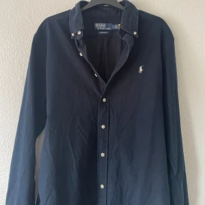 Mörkblå skjorta från Polo Ralph Lauren - Klassisk mörkblå skjorta från Polo Ralph Lauren i bomull med custom fit. Skjortan har långa ärmar, knappar framtill och den ikoniska broderade loggan på bröstet. Perfekt för dig som gillar stilren och tidlös design.