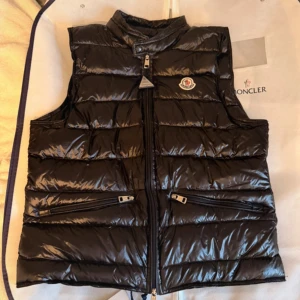 Svart dunväst från Moncler - Snygg svart dunväst från Moncler med glansig finish och klassisk logga på bröstet. Västen har hög krage, två dragkedjefickor framtill och stängs med dragkedja. Perfekt att slänga över en hoodie för en cool look.