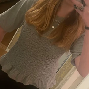 Grå smockad topp med volangkant - Supersöt grå topp med smockad midja och volangkant nertill. T-shirtrmar och rund halsringning ger en avslappnad vibe. Perfekt att styla med jeans eller kjol för en chill look.