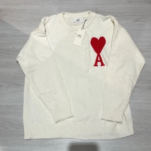 Vit stickad tröja med rött hjärta från Ami - Vit stickad tröja från Ami Alexandre Mattiussi med ett stort rött hjärta och bokstaven A på bröstet. Tröjan har rund hals, ribbade muddar och är tillverkad av ekologisk bomull. Perfekt för dig som gillar stilrena plagg med en twist.