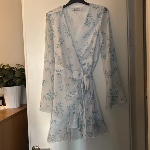 Blommig omlottklänning i ljusblått - Superfin kort omlottklänning med blommigt mönster i ljusblå färg. Klänningen har långa ärmar, v-ringning och volangdetalj framtill. Perfekt för sommarens alla tillfällen. 