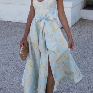 Ljusblå blommig långklänning med volang - Superfin ljusblå långklänning med blommigt mönster i vitt och beige. Klänningen har volangdetaljer på axelbanden, markerad midja med knytband och en snygg slits framtill. Perfekt för sommarens alla tillfällen, till exempel bröllopsgäst
