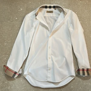 Vit skjorta från Burberry med rutiga detaljer - Vit skjorta från Burberry med klassisk krage och långa ärmar. Ikoniska rutiga detaljer syns på insidan av kragen och ärmsluten. Skjortan har knäppning framtill och är tillverkad i bomull för en skön och stilren känsla.