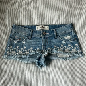 Blå broderade jeansshorts Hollister - Supersnygga blå jeansshorts från Hollister med slitna detaljer, fransig kant och vit blommig brodyr längs benen. Midjemått ca 35 cm rakt över, grenhöjd ca 16,5 cm, längd ca 17 cm 💘