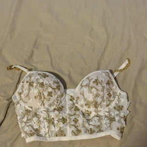 Vit och guldig broderad bralette - Superfin vit bralette med broderade blommor i guld och vita toner. Axelbanden är delvis i guldfärgad kedja och delvis justerbara band. Kuporna är vadderade och toppen har hakknäppning i ryggen. Från SHEIN och i storlek i S, har använt den endast en gång. 