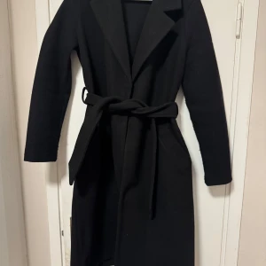 Svart kappa med bälte från Vero Moda - Stilren svart kappa från Vero Moda i klassisk modell med bälte i midjan och långa ärmar. Snygg krage och djupa fickor framtill. 