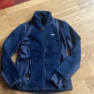 Marinblå teddyjacka från JH Collection - Snygg marinblå teddyjacka från JH Collection med mjuk, fluffig yta och coola vågiga detaljer. Jackan har hel dragkedja framtill, två fickor med dragkedja och hög krage. Perfekt för dig som gillar stilren och sportig look med extra komfort.