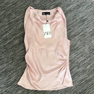 Ljusrosa satinlinnetopp från Zara - Supersnygg ljusrosa satintopp från Zara. Toppen har en draperad halsringning och är ärmlös. Aldrig använd med prislappen kvar❤️ pris kan diskuteras!
