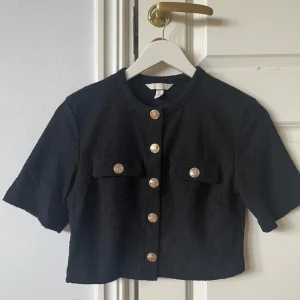 Svart croppad blus med guldknappar H&M - Stilren svart croppad blus från H&M med korta ärmar och snygga guldknappar framtill. Toppen har två design fickor med guld knappar och struktur i tyget som ger en lyxig känsla. Perfekt för dig som gillar trendiga detaljer och klassisk look.