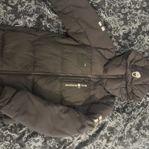 Svart pufferjacka Sail Racing - Svart pufferjacka från Sail Racing med huva och dragkedja framtill. Jackan har broderad logga på bröstet och ärmarna, samt en quiltad design som ger en schysst streetkänsla. Perfekt för kalla dagar när du vill hålla stilen. Köpt 2024