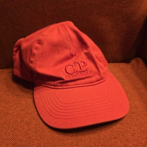 Röd C.P. Company keps med goggles - Säljer en röd keps från C.P. Company med deras ikoniska goggles bak. Kepsen har böjd skärm, broderad logga framtill och är tillverkad i bomull. Perfekt för dig som vill sticka ut med en streetwear-inspirerad look.