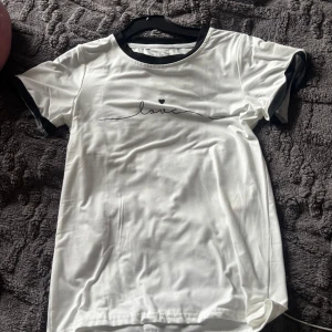 Vit t-shirt med svart text och detaljer - Vit t-shirt med svarta detaljer vid halsringning och ärmslut. Tryck med ordet 'love' och ett litet hjärta på bröstet. Klassisk passform och korta ärmar, perfekt för en chill och stilren look.