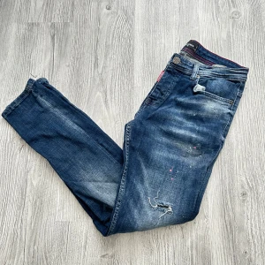 Dsquared2 blå jeans med slitningar - Snygga blå jeans från Dsquared2 med coola slitningar och färgstänk. Jeansen har en klassisk femficksmodell, röd detaljsöm och Dsquared2-lapp bak. Perfekt för dig som gillar en edgy streetstil.