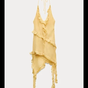 Gul asymmetrisk klänning från Zara - Supersöt gul kortklänning från Zara med volangdetaljer och asymmetrisk nederkant. Klänningen har smala axelband som knyts i nacken och v-ringning framtill. Lätt och flowigt material, perfekt för varma dagar.