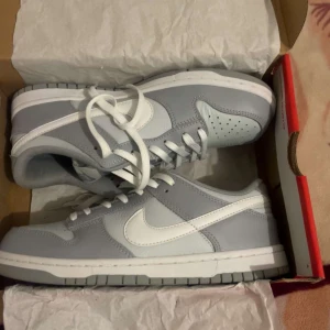 Nike Dunk Low i grått och vitt - Nike Dunk Low sneakers i stilren grå och vit färg. Skorna har klassisk låg siluett, vit sula och snörning. Tillverkade i skinn med mjuk vadderad plös och ikonisk Swoosh-logga på sidorna. Perfekta för dig som gillar clean och modern streetstyle.