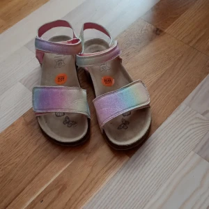 Skin skor Glittriga regnbågsfärgade sandaler  - Säljer ett par sandaler i regnbågsfärgat, glittrigt material med kardborreband. Skorna har platt sula och öppen tå, perfekt för varma dagar. Insidan är mjuk och dekorerad med fjärilar. Färgerna skiftar i rosa, lila, blått och gult.