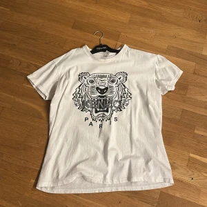 Vit Kenzo t-shirt med tigertryck - Snygg vit t-shirt från Kenzo med ikoniskt svart tigertryck och texten 'Kenzo Paris' framtill. Klassisk passform och rund halsringning, perfekt för en cool och avslappnad stil.