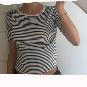 Randig ribbad topp med kort ärm - Säljer en vit och svart randig ribbad topp med korta ärmar och rund halsringning. Toppen har en croppad passform och vågig kant nertill för en extra snygg detalj. Perfekt till jeans eller kjol för en chill look.