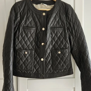 Svart quiltad skinnjacka DAY - Snygg svart quiltad skinnjacka från DAY Birger et Mikkelsen. Jackan har rund hals, guldiga knappar framtill och två fickor med lock. Klassisk och stilren modell med lång ärm och rutmönstrad yta. Perfekt för dig som gillar en clean och trendig look.