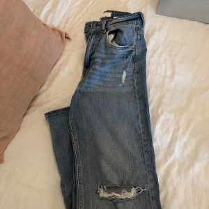 Blå slitna jeans från H&M, strl 36 - Säljer ett par blå jeans från H&M i storlek 36. Jeansen har en klassisk femficksmodell, slitningar på låret och knäet för en cool, avslappnad look. Tillverkade i bomull med raka ben och normal passform.