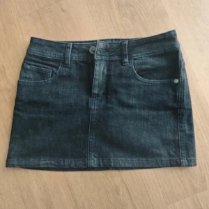 Svart mini jeanskjol från Snazee - Snygg mörkblå jeanskjol från Snazee med klassisk femficksdesign, bälteshällor och knappstängning fram. Kjolen är kort o snygg midjan är 34 cm o total längden 25