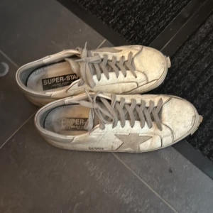 Golden Goose Superstar sneakers beige - Golden Goose Superstar sneakers i beige med grå stjärndetalj på sidan och grå snörning. Klassisk låg modell med platt sula och rund tå. Skorna har GGDB-logga och är tillverkade i skinn för en cool och avslappnad look. Som sagt är skorna väldigt använda och därefter är priset så lågt!!
