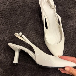 Vita klackskor från Bershka - Snygga vita klackskor från Bershka med spetsig tå och smal klack. Skorna har öppen häl med justerbart spänne och är tillverkade i glansigt syntetmaterial. Perfekta för dig som vill sticka ut med en stilren och trendig look.