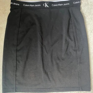 Svart kortkjol från Calvin Klein Jeans - Svart kortkjol från Calvin Klein Jeans med elastisk midja och logotypband runt hela linningen. Enkel och clean design som är lätt att matcha med olika toppar. Kjolen har en rak passform och är tillverkad i mjukt material för en bekväm känsla. Kjolen är i storlek 16 men passar XS/S