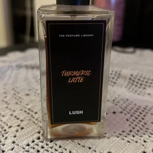 Lush Turmeric Latte Parfym 100ml - Unik parfym från Lush, Turmeric Latte, i en stilren, fyrkantig glasflaska med svart etikett och lock. Doften har mjuka toner av vanilj, mjölk och tonkaböna, blandat med kryddig gurkmeja. Innehåller eteriska oljor och är vegansk. 100 ml. 85% kvar 