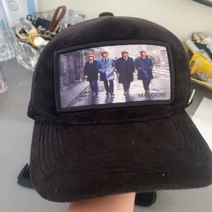 Svart keps med Maggiore-tryck - Svart keps från Maggiore med mesh på sidorna och ett stort tygmärke framtill som visar fyra personer i kostym på en gata. Kepsen har böjd skärm och cool streetstyle-vibe, perfekt för att sticka ut i mängden. Har använt den runt 4-5 gånger den är i bra qualite och har inga fläckar.