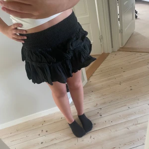 Svart volangkjol från Blanco by Natural - Supersöt svart kortkjol med volanger från Blanco by Natural. Kjolen har short under så man inte flashat. Kjolen har bred smock i midjan för en bekväm passform och är gjord i ett lätt, skrynkligt tyg som ger en cool och avslappnad vibe. Perfekt att styla med crop top eller hoodie.