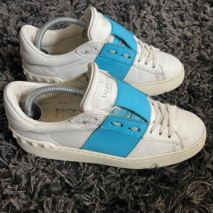 Valentino skor - Snygga vita sneakers från Valentino Garavani med en bred blå läderdetalj över snörningen. Skorna har rund tå, platt sula och klassisk snörning. Tillverkade i skinn med coola nitar på hälen för extra edge. Perfekta för dig som gillar statement sneakers.