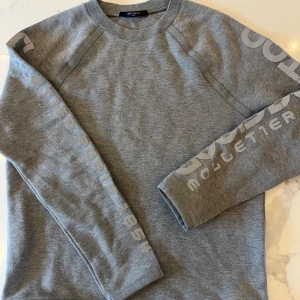 Grå sweatshirt från Louis Vuitton - Snygg grå sweatshirt från Louis Vuitton med tryckt logga längs ärmarna. Tröjan har rund halsringning, ribbade muddar och en clean, stilren look. Perfekt för dig som gillar exklusiva streetwear-vibbar och vill sticka ut med ett lyxigt märke. 