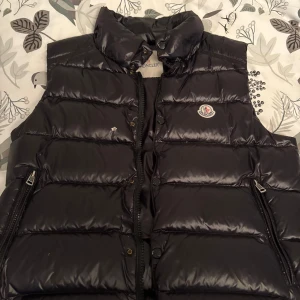 Svart dunväst från Moncler - Snygg svart dunväst från Moncler med hög krage och klassisk logga på bröstet. Västen har blanka paneler, dragkedja framtill och två fickor med dragkedja. Perfekt för lager-på-lager och ger en clean, sportig look.
