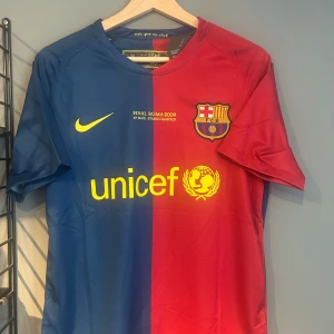 FC Barcelona Messi 10 Final Roma 2009 tröja - Säljer en ikonisk FC Barcelona fotbollströja från Final Roma 2009 med Messi 10 på ryggen. Tröjan är blå och röd med gula detaljer, klubbmärke och Nike-logga framtill samt UNICEF-tryck. Tillverkad i lätt och ventilerande material, perfekt för fans. Helt ny med tagg en replika 