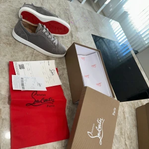 Grå Christian Louboutin skor - Snygga grå sneakers i mocka från Christian Louboutin med klassisk röd sula och vita kanter. Skorna har rund tå, snörning och diskreta sömdetaljer. Perfekta för dig som vill ha en stilren och lyxig look med en ikonisk touch.
