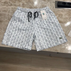 Lacoste Badshorts - De är storlek L men passar även M. Snygga vita badshorts från Lacoste med blått geometriskt mönster och svart snörning i midjan. Klassisk passform och broderad grön krokodil på benet. Perfekta för sommardagar vid poolen eller stranden.