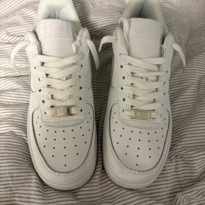 Nike Air Force 1 vita sneakers - Klassiska Nike Air Force 1 sneakers i helvitt skinn med perforerad tå, rund tå och platt sula. Skorna har snörning med vita skosnören och metallbricka med 'AF1' framtill. Perfekt för dig som gillar en clean och tidlös look. Helt nya inte använt en enda GÅNG.