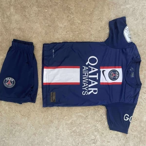 PSG Neymar Jr matchtröja set - Paris Saint-Germain fotbollströja och shorts med Neymar Jr och nummer 10 på ryggen. Tröjan är marinblå med vit och röd vertikal rand, PSG-logga och Qatar Airways-tryck. Tillverkad i lätt och ventilerande material, perfekt för fotbollsträning eller match.