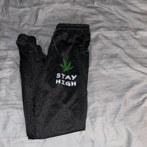 Svarta mjukisbyxor med tryck - Mörkgrå mjukisbyxor med resår och snörning i midjan. På ena benet finns ett grönt blad och texten 'STAY HIGH' i vitt. Byxorna har avslappnad passform och är perfekta för chill dagar.