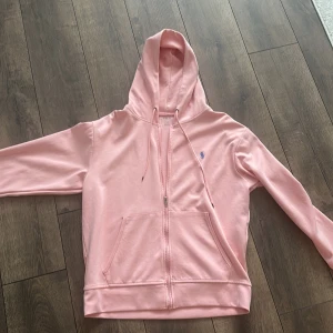 Ljusrosa hoodie från Polo Ralph Lauren - Säljer en ljusrosa hoodie från Polo Ralph Lauren med dragkedja och huva. Tröjan har snörning i huvan, känguruficka och blå broderad logga på bröstet. Tillverkad i mjuk bomull, perfekt för chill dagar. Klassisk och stilren design.
